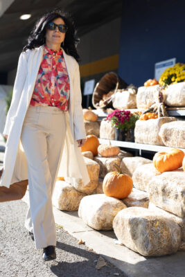 Model wearing walks by stand decorated with straw bales and pumpkins wearing white cardigan, greige pants and floral print blouse