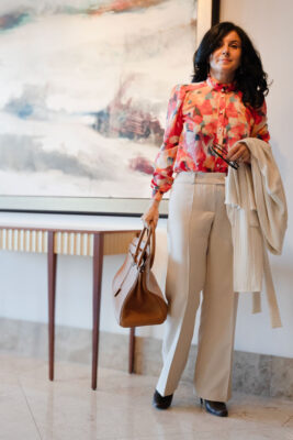 Model standing next to a table wearing greige pants and printed blouse and holding a white cardigan over her hand