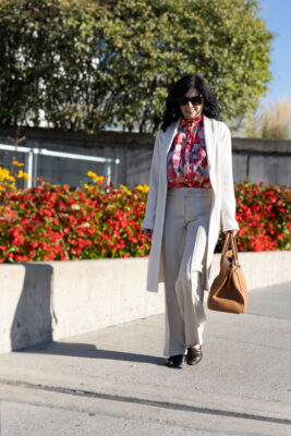 Model walking on sidewalk wearing white cardigan and pants and a printed blouse