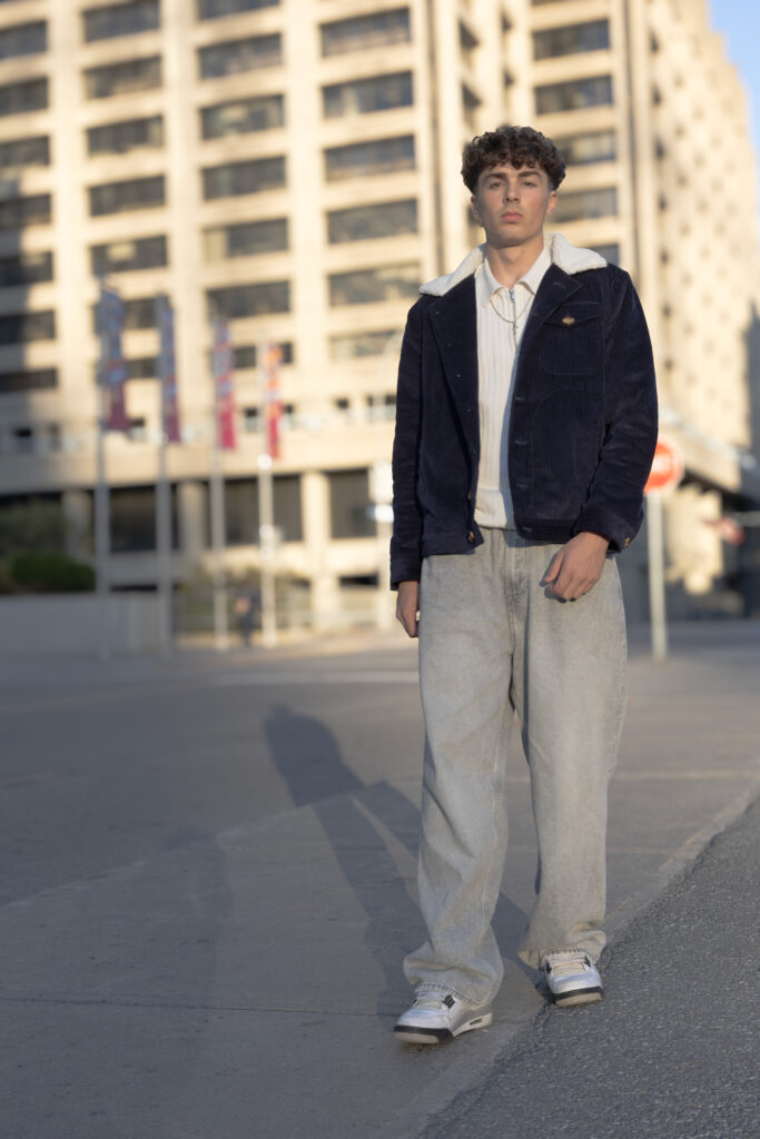 model wearing navy corduroy jacket with white faux fur lining, grey jeans, white polo and white sneakers walking down a street with high rise office buildings in the background