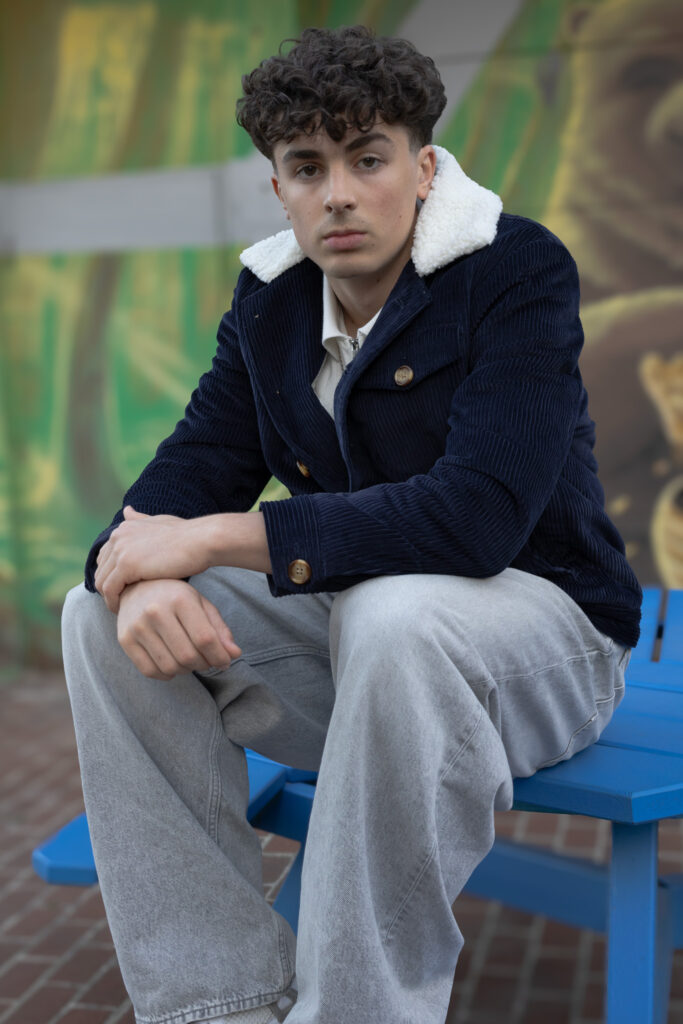 model wearing navy corduroy jacket with white faux fur lining and grey jeans sits on a blue table